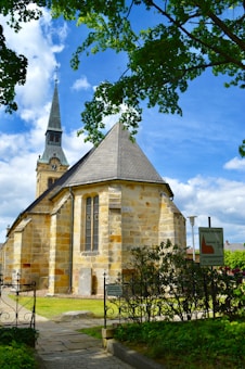 A historic stone church with a tall spire rises against a backdrop of a bright blue sky with scattered clouds. The church is surrounded by lush green trees and bushes, and in the foreground, decorative wrought iron fencing lines a pathway. A sign next to the fence indicates some information in German.