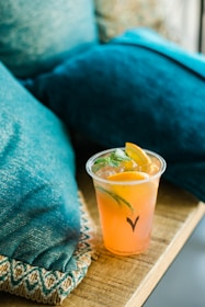 A refreshing drink with slices of lemon and lime in a clear plastic cup sits on a wooden table. The setting includes textured turquoise pillows, suggesting a casual and cozy environment.
