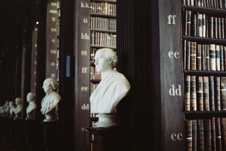 The image depicts a serene library scene with two white busts of prominent figures, one on each side of a row of bookshelves filled with numerous books. The busts are positioned at different angles and heights, creating an interesting visual dynamic within the space. The background is dark, providing a stark contrast to the lighter elements in the foreground.