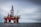 An offshore oil rig standing prominently in the middle of the ocean, featuring red and metallic structures with cranes attached. The platform is supported by large cylindrical legs, and the sea stretches out beneath a cloudy sky. A small boat is visible near the base of the rig, emphasizing its massive scale.