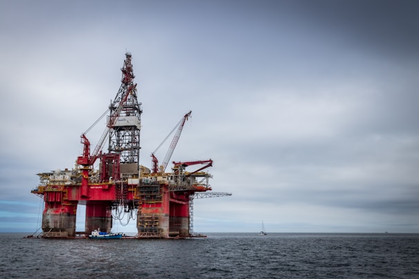 An offshore oil rig standing prominently in the middle of the ocean, featuring red and metallic structures with cranes attached. The platform is supported by large cylindrical legs, and the sea stretches out beneath a cloudy sky. A small boat is visible near the base of the rig, emphasizing its massive scale.
