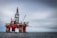 An offshore oil rig standing prominently in the middle of the ocean, featuring red and metallic structures with cranes attached. The platform is supported by large cylindrical legs, and the sea stretches out beneath a cloudy sky. A small boat is visible near the base of the rig, emphasizing its massive scale.