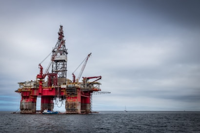 An offshore oil rig standing prominently in the middle of the ocean, featuring red and metallic structures with cranes attached. The platform is supported by large cylindrical legs, and the sea stretches out beneath a cloudy sky. A small boat is visible near the base of the rig, emphasizing its massive scale.