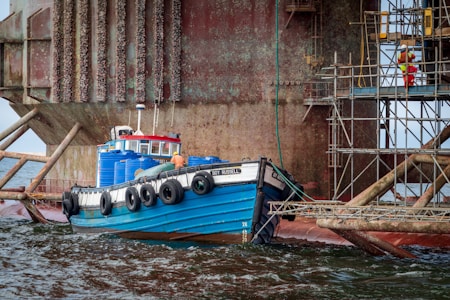 A small blue and black tugboat is docked next to an industrial structure at sea. The boat is equipped with large black fenders along its side, and several blue barrels are stacked on its deck. The background features a weathered steel structure with barnacles, scaffoldings, and a person wearing bright red and yellow protective clothing climbing it.