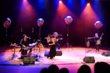 A musical performance on stage with four musicians. One plays the drums, another is on a guitar, the third is a vocalist holding a microphone, and the fourth plays a string instrument similar to a banjo. They are surrounded by large stage lights that cast a mix of blue, purple, and orange hues, creating a vibrant and lively atmosphere.