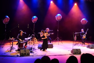 A musical performance on stage with four musicians. One plays the drums, another is on a guitar, the third is a vocalist holding a microphone, and the fourth plays a string instrument similar to a banjo. They are surrounded by large stage lights that cast a mix of blue, purple, and orange hues, creating a vibrant and lively atmosphere.