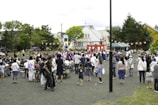 A community festival is taking place in an open space with a crowd of people, including adults and children, gathered around a central raised platform adorned with red and white stripes. Many attendees are dressed in traditional clothing, such as kimonos. A string of red paper lanterns hangs in the background, and some people are seated while others stand and engage in various activities. Trees and residential buildings are visible in the background.