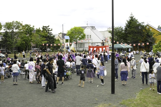 A community festival is taking place in an open space with a crowd of people, including adults and children, gathered around a central raised platform adorned with red and white stripes. Many attendees are dressed in traditional clothing, such as kimonos. A string of red paper lanterns hangs in the background, and some people are seated while others stand and engage in various activities. Trees and residential buildings are visible in the background.