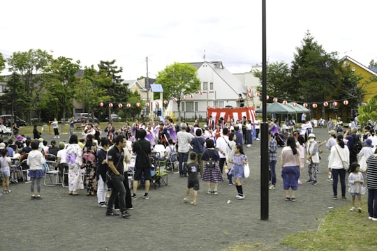 A community festival is taking place in an open space with a crowd of people, including adults and children, gathered around a central raised platform adorned with red and white stripes. Many attendees are dressed in traditional clothing, such as kimonos. A string of red paper lanterns hangs in the background, and some people are seated while others stand and engage in various activities. Trees and residential buildings are visible in the background.