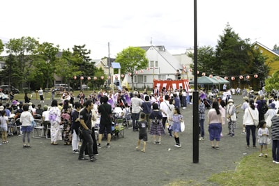 A community festival is taking place in an open space with a crowd of people, including adults and children, gathered around a central raised platform adorned with red and white stripes. Many attendees are dressed in traditional clothing, such as kimonos. A string of red paper lanterns hangs in the background, and some people are seated while others stand and engage in various activities. Trees and residential buildings are visible in the background.