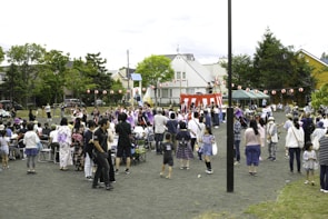 A community festival is taking place in an open space with a crowd of people, including adults and children, gathered around a central raised platform adorned with red and white stripes. Many attendees are dressed in traditional clothing, such as kimonos. A string of red paper lanterns hangs in the background, and some people are seated while others stand and engage in various activities. Trees and residential buildings are visible in the background.