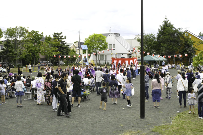 A community festival is taking place in an open space with a crowd of people, including adults and children, gathered around a central raised platform adorned with red and white stripes. Many attendees are dressed in traditional clothing, such as kimonos. A string of red paper lanterns hangs in the background, and some people are seated while others stand and engage in various activities. Trees and residential buildings are visible in the background.