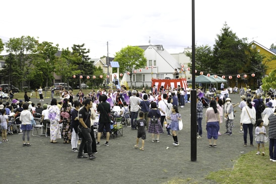 A community festival is taking place in an open space with a crowd of people, including adults and children, gathered around a central raised platform adorned with red and white stripes. Many attendees are dressed in traditional clothing, such as kimonos. A string of red paper lanterns hangs in the background, and some people are seated while others stand and engage in various activities. Trees and residential buildings are visible in the background.