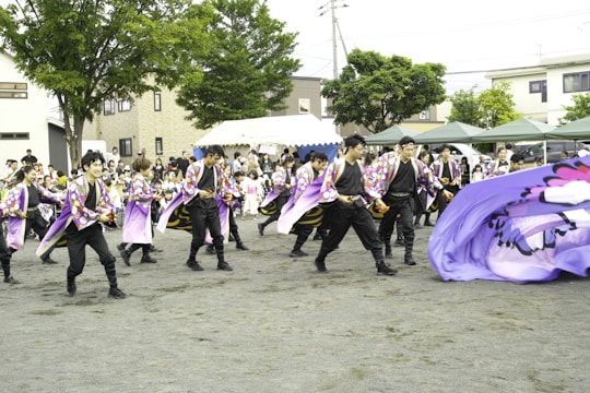 A group of performers, dressed in vibrant purple and black traditional attire, are engaged in a coordinated dance. They are in an outdoor setting with a crowd of spectators seated under tents and trees in the background.