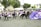 A group of performers, dressed in vibrant purple and black traditional attire, are engaged in a coordinated dance. They are in an outdoor setting with a crowd of spectators seated under tents and trees in the background.