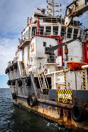 A large rescue ship on the water is visible, featuring a robust structure with multiple decks and safety equipment on board. The hull is painted with a combination of white and dark colors, and there are several large tires attached to the side, presumably for protection or docking purposes. Red pipes and lifebuoys are mounted on the exterior, and the words 'RESCUE' and 'SAFETY FIRST' are prominently displayed. The ship is equipped with cranes and other machinery, indicating its readiness for emergency situations.