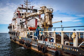 A large industrial ship is on the water, equipped with various machinery and safety equipment. The ship has a prominent crane and various piping and railing structures, with lifeboats and tires along the side for docking. The name 'Mamola Challenger Kingston' is visible on the side. The sky above is a mix of clouds and clear blue.