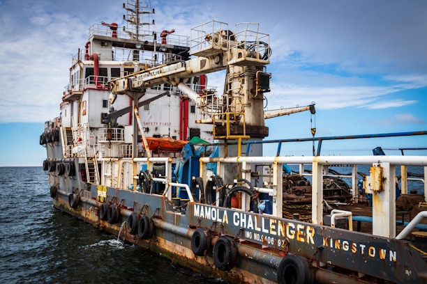 A large industrial ship is on the water, equipped with various machinery and safety equipment. The ship has a prominent crane and various piping and railing structures, with lifeboats and tires along the side for docking. The name 'Mamola Challenger Kingston' is visible on the side. The sky above is a mix of clouds and clear blue.