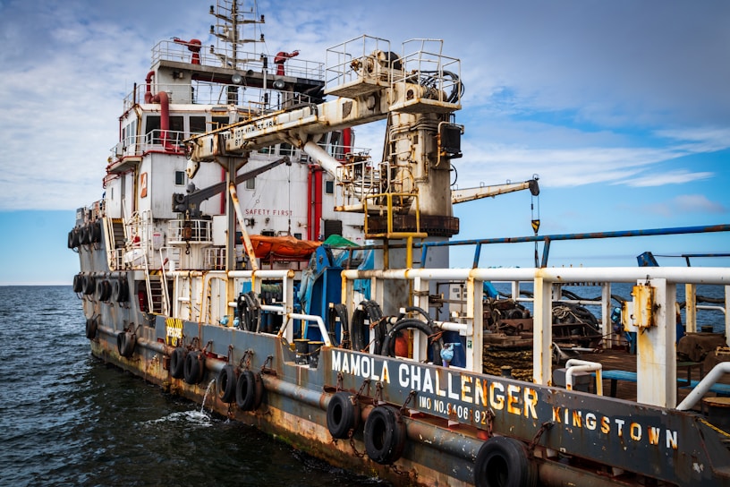 A large industrial ship is on the water, equipped with various machinery and safety equipment. The ship has a prominent crane and various piping and railing structures, with lifeboats and tires along the side for docking. The name 'Mamola Challenger Kingston' is visible on the side. The sky above is a mix of clouds and clear blue.