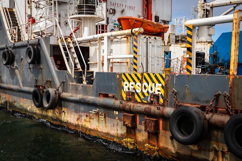 A close-up view of a ship's side featuring various safety equipment and details. Prominent black and yellow striped 'RESCUE' signage is visible, along with several attached tire bumpers used as fenders. The metal structure displays signs of rust and wear, and various stairs and railings are part of the ship's infrastructure. An orange lifeboat is prominently visible in the upper section.
