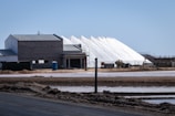 Several large white pyramidal mounds stand near industrial buildings under a clear blue sky. The area appears to be a salt processing facility, with noticeable mounds of salt and construction equipment scattered around.