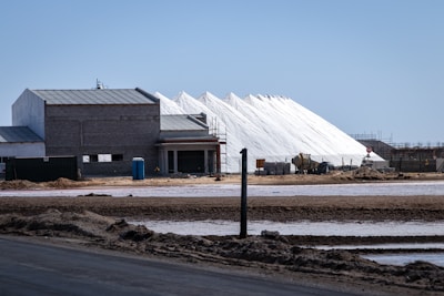 Several large white pyramidal mounds stand near industrial buildings under a clear blue sky. The area appears to be a salt processing facility, with noticeable mounds of salt and construction equipment scattered around.