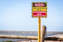 A warning sign is prominently displayed, attached to a yellow post with a chain. The sign indicates a mine lease area, ML 37, with a message that no unauthorized entry is allowed and trespassers will be prosecuted. The sign is red and yellow with black stripes, positioned in an open, natural landscape with blue sky above.