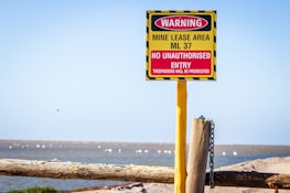 A warning sign is prominently displayed, attached to a yellow post with a chain. The sign indicates a mine lease area, ML 37, with a message that no unauthorized entry is allowed and trespassers will be prosecuted. The sign is red and yellow with black stripes, positioned in an open, natural landscape with blue sky above.