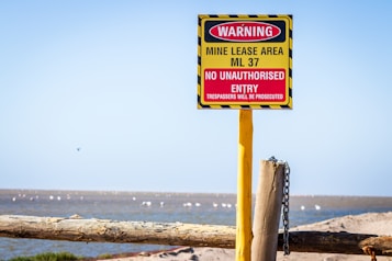 A warning sign is prominently displayed, attached to a yellow post with a chain. The sign indicates a mine lease area, ML 37, with a message that no unauthorized entry is allowed and trespassers will be prosecuted. The sign is red and yellow with black stripes, positioned in an open, natural landscape with blue sky above.