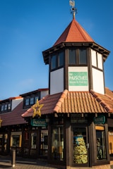A quaint building with traditional architecture featuring a pointed red-tiled roof and a decorative weather vane on top. The storefront has large windows and displays a sign for 'Die Muschel Books & Fine Art.' Bright yellow star decorations are mounted on the facade, adding a festive touch. The surrounding area looks well-maintained, with cobblestone walkways.