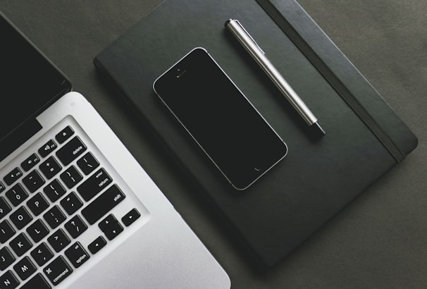 A close-up view of a laptop keyboard, a smartphone, a closed black notebook, and a silver pen arranged on a gray surface. The laptop is partially shown with its keyboard illuminated. The smartphone is placed on top of the notebook next to the pen.