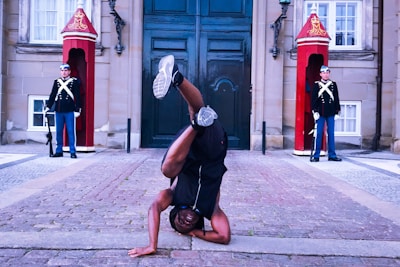 A person performs a one-handed handstand on a cobbled pathway in front of a historical building with two guards dressed in formal uniforms standing in front of red guardhouses on either side.