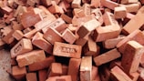 A pile of red clay bricks, some of which have text imprints. The bricks are stacked unevenly and are slightly weathered.