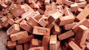 A pile of red clay bricks, some of which have text imprints. The bricks are stacked unevenly and are slightly weathered.