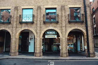 A brick building with three arched doorways, each leading to different areas. The middle archway features a coffee shop named 'Mangio Coffee'. The upper floor includes three windows with wrought iron railings. Several elements of a cozy urban setting are visible, including a paved street in front.