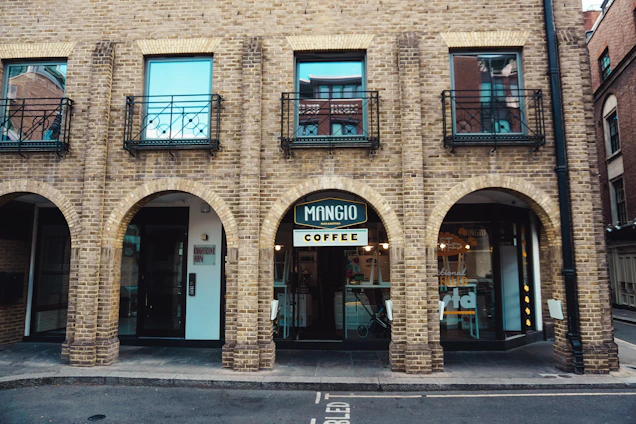 A brick building with three arched doorways, each leading to different areas. The middle archway features a coffee shop named 'Mangio Coffee'. The upper floor includes three windows with wrought iron railings. Several elements of a cozy urban setting are visible, including a paved street in front.