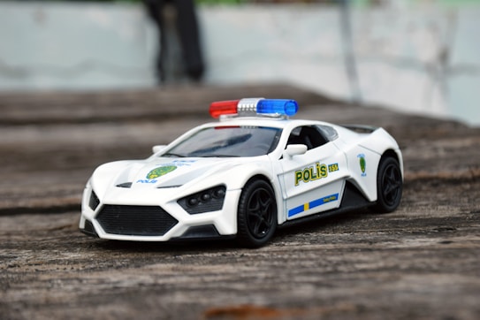 A detailed toy police car with a sleek, sports car design is placed on a textured wooden surface. The car is primarily white, featuring police decals and a light bar with red and blue lights on the roof. The wheels and windows are black, adding contrast to the overall appearance.