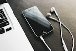 A smartphone lies on a dark surface next to a pair of white earphones, with part of a laptop keyboard visible in the corner.