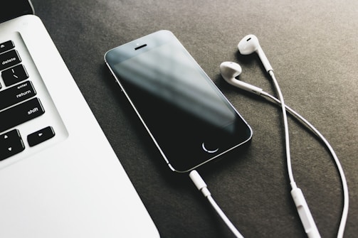 A smartphone lies on a dark surface next to a pair of white earphones, with part of a laptop keyboard visible in the corner.