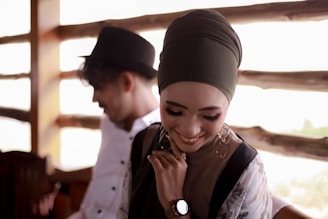 A woman wearing a hijab is smiling and looking down, appearing joyful and content. She is accessorized with earrings and a watch, and appears to be dressed in a stylish manner. In the background, a man wearing a hat is slightly blurred, adding depth to the composition. The setting seems to be a cozy, rustic wooden interior.