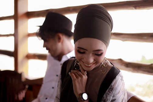 A woman wearing a hijab is smiling and looking down, appearing joyful and content. She is accessorized with earrings and a watch, and appears to be dressed in a stylish manner. In the background, a man wearing a hat is slightly blurred, adding depth to the composition. The setting seems to be a cozy, rustic wooden interior.