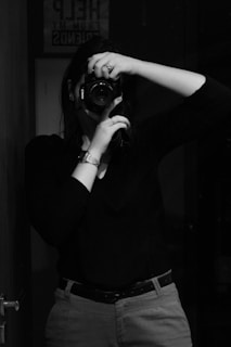 A person is holding a camera up to their face, capturing a self-portrait in a mirror. They are wearing a dark long-sleeved shirt and light-colored pants with a belt. The room is dimly lit, creating a high-contrast, monochrome effect. A framed sign with partial text is visible in the background.
