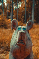A carved wooden bear head wearing blue-tinted glasses stands against a blurred forest background. The forest features tall trees and autumn-colored foliage, creating a warm, rustic atmosphere.
