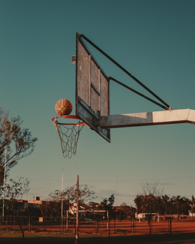 A basketball hoop with a net attached stands prominently against a clear sky as a basketball is caught mid-air, about to go through the hoop. In the background, trees and a fence are visible, illustrating an outdoor sports area.