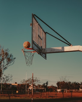 A basketball hoop with a net attached stands prominently against a clear sky as a basketball is caught mid-air, about to go through the hoop. In the background, trees and a fence are visible, illustrating an outdoor sports area.