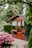 A small wooden garden shed with a glass door, flanked by windows with lace curtains. Red and purple flowers in pots are placed outside the windows. Surrounding the shed are well-maintained hedges, large trees, and a vibrant bush with pink flowers. A paved pathway leads to the shed.