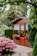 A small wooden garden shed with a glass door, flanked by windows with lace curtains. Red and purple flowers in pots are placed outside the windows. Surrounding the shed are well-maintained hedges, large trees, and a vibrant bush with pink flowers. A paved pathway leads to the shed.