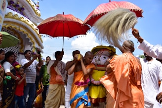 A vibrant outdoor scene featuring a group of people engaged in a traditional ceremony. Several individuals are wearing orange robes, holding a large decorated red umbrella. Others carry ceremonial items and fan with traditional whisk-like objects. A large colorful idol or religious figure is in the center, surrounded by participants. The backdrop includes ornate temple architecture with intricate designs.