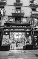 An exterior view of an old pharmacy building with vintage signage. The entrance features large glass doors displaying the word 'pharmacy' and posters on either side. The building has multiple storeys with balconies on the upper levels.