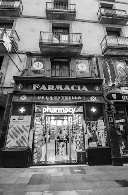 An exterior view of an old pharmacy building with vintage signage. The entrance features large glass doors displaying the word 'pharmacy' and posters on either side. The building has multiple storeys with balconies on the upper levels.